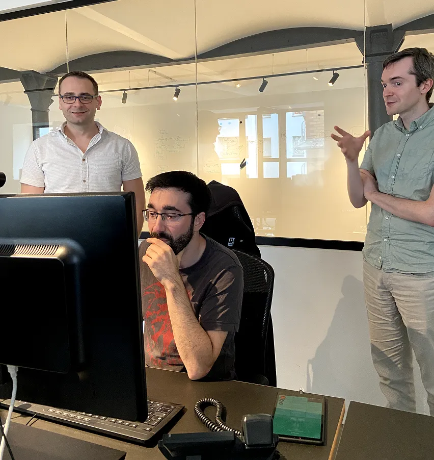 Martin steht mit zwei Kollegen im Büro vor einem Bildschirm und diskutiert eine technische Aufgabe. Konzentriertes Teamwork im Development-Alltag.