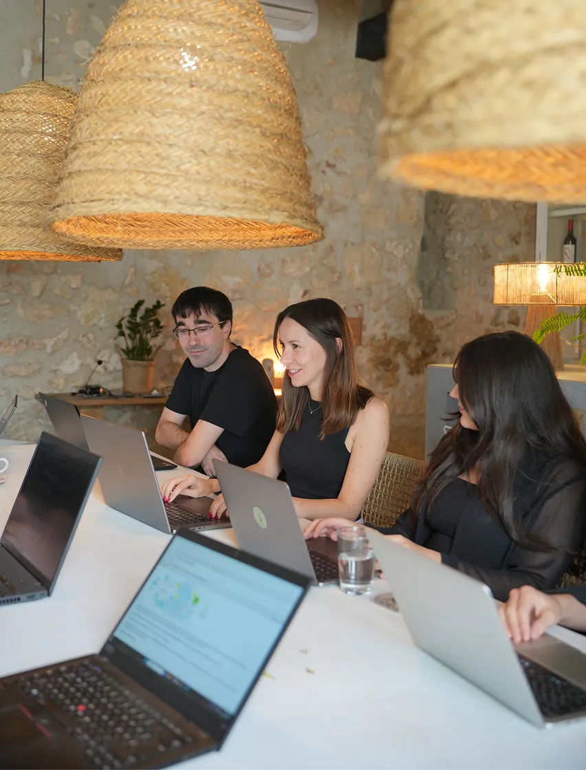 Mehrere Personen sitzen an einem langen Tisch in einem hellen Raum mit Natursteinwänden und großen geflochtenen Lampen. Sie arbeiten gemeinsam an Laptops und wirken konzentriert. Die Szene zeigt kollaboratives Remote-Teamwork.
