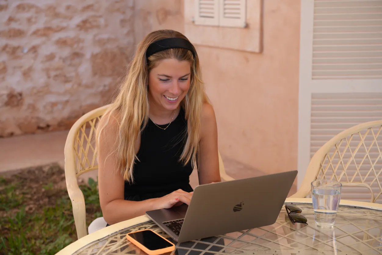 Theresa sitzt mit Laptop im Freien, fokussiert auf den Bildschirm. Ein sonniger Arbeitstag in mediterraner Umgebung bei der Workation.